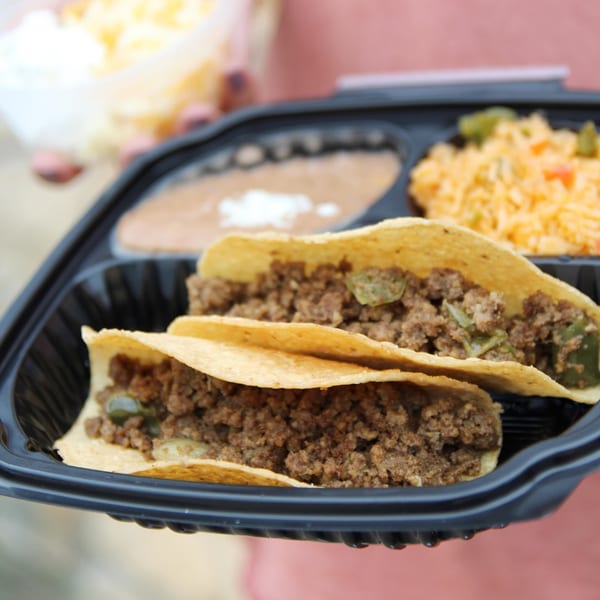 Two ground beef tacos, refried beans, and Mexican rice in a black takeout container.