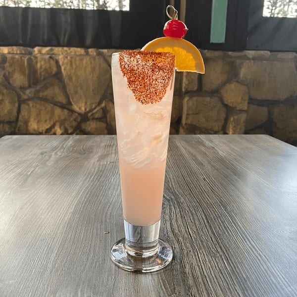 Pink cocktail with a chili-lime rim, orange slice, and cherry garnish on a wooden table.